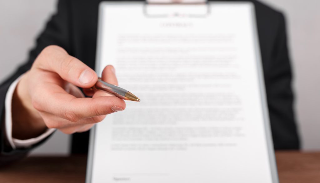 Businessman handing over a contract, attached to a clipboard for signature, offering a ballpoint pen in his hand.