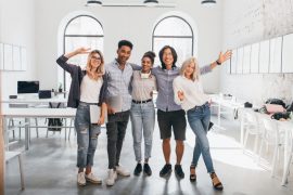 Full-length portrait of slim female office worker in jeans standing with legs crossed near asian colleague. Indoor photo of tall african student and glad european girl with laptop..