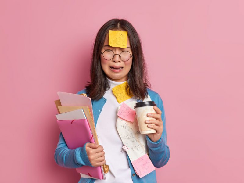 Overworked frustrated female accountant has much remote work surrounded with paper documents drinks coffee to go cries from despair isolated over pink background. Upset woman employee has problems